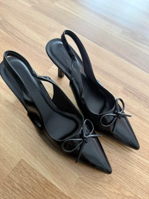 Svarta lackpumps med rosett - Snygga svarta pumps i lack med spetsig tå och smal klack. Skorna har öppen häl med slingback-rem och en söt rosett framtill. Perfekta för dig som vill ha en elegant och trendig look.