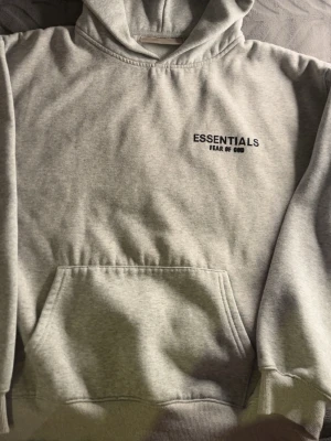Grå Essentials Fear of God hoodie - Snygg grå hoodie från Fear of God Essentials med svart logga på bröstet och ryggen. Klassisk känguruficka framtill och huva. Perfekt för en avslappnad streetwear-look. Mjuk bomullsblandning som känns skön mot huden.