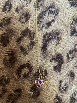 Halsband med lila hjärthänge och pil guld - Guldfärgat halsband med ett hjärthänge i lila sten, inramat av en pil i metall. Kedjan är tunn och elegant, och hänget har en lekfull och romantisk design. Perfekt accessoar för att lyfta din outfit med en färgklick och unik detalj. Rostfri 