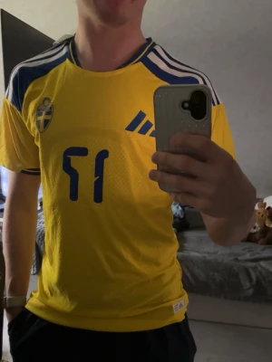Svensk landslagströja Adidas gul, Viktor Gyökeres  - Gul svensk landslagströja från Adidas med blå detaljer och nummer 17 på bröstet. Tröjan har korta ärmar, blå-vita ränder på axlarna och Sveriges landslagsemblem på vänster sida. Perfekt för fotbollsfans som vill visa sitt stöd.