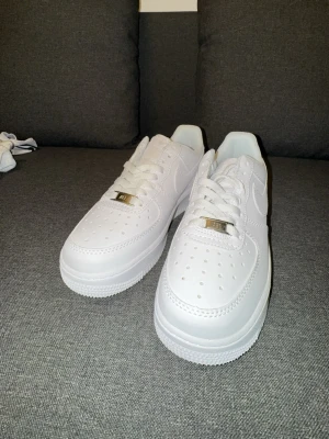 Nike Air Force 1 vita sneakers - Klassiska Nike Air Force 1 sneakers i helvitt läder med perforerad tå, vit sula och vita skosnören. Snygg metallbricka med 'AF1' på snörningen och ikonisk Nike Swoosh på sidorna. Perfekt för dig som gillar en clean och tidlös look.