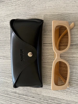 Beige fyrkantiga solglasögon från SHEIN - Snygga solglasögon från SHEIN med beige, fyrkantig båge och trendig retrostil. Kommer med svart fodral i syntetmaterial. Perfekta för att lyfta din look under soliga dagar.