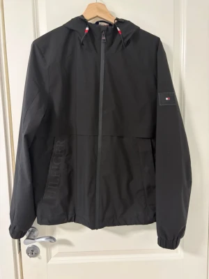 Svart vindjacka från Tommy Hilfiger - Snygg svart vindjacka från Tommy Hilfiger med huva och dragkedja framtill. Jackan har diskret Hilfiger-logga på vänster ärm och broderad text på fickan. Elastiska muddar vid ärmslut och justerbar huva med röda och vita detaljer på snöret.