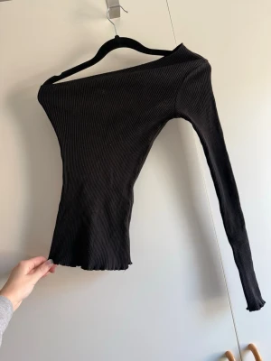 Svart ribbad oneshoulder topp - Trendig svart ribbad topp med oneshoulder-design och lång ärm. Materialet är stretchigt och följsamt, vilket ger en slim och snygg passform. Perfekt för dig som vill sticka ut med en unik och stilren look. Köpt begagnad, obs katt finns i hem!