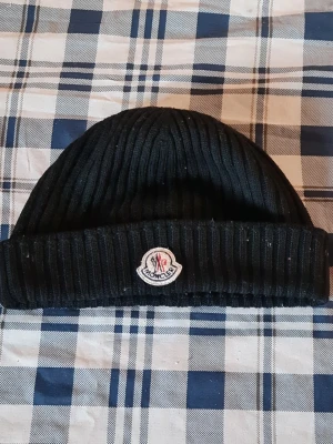 Svart Moncler mössa i ribbstickad ull - Snygg svart ribbstickad mössa från Moncler med klassisk uppvikt kant och broderad logotyp framtill. Tillverkad i mjuk ull som håller dig varm och ger en stilren look. Perfekt accessoar för kalla dagar och streetwear-stil.
