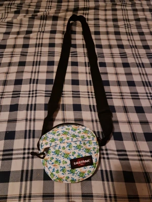Blommig rund väska från Eastpak - Säljer en liten rund väska från Eastpak med blommigt mönster i grönt, blått och gult på vit bakgrund. Väskan har en svart justerbar axelrem och dragkedja. Perfekt för att bära småsaker och ge din outfit en färgglad touch.