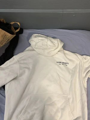 Vit hoodie Future Projects NYC - Vit hoodie från Future Projects med svart tryck på bröstet och stor text på ryggen: 'Future Projects New York City'. Har huva och magficka. Snygg och enkel design, perfekt för dig som gillar streetwear och vill sticka ut med en clean look.