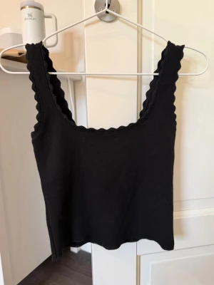 Svart ribbad linnetopp med volangkant - Svart linnetopp med breda axelband och vågig volangkant längs ringningen och axelbanden. Toppen är ribbad och har en croppad passform, perfekt att styla till jeans eller kjol. Materialet känns mjukt.