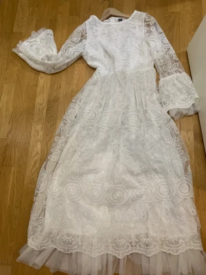 Vit lång spetsklänning med volangärm - Romantisk vit långklänning med vackra spetsdetaljer och broderat mönster. Klänningen har långa, vida ärmar med volang och transparenta partier samt en mjuk tyllkant nertill. Perfekt för speciella tillfällen och ger en drömmig känsla.