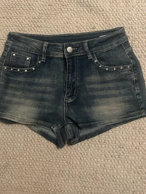 SHEIN Jeansshorts med döskallebrodyr - Mörkblå jeansshorts från SHEIN med slitningar och coola döskallebroderier på bakfickorna. Framtill finns nitar på fickorna och klassisk knappstängning. Shortsen har en slim passform och är perfekta för dig som vill sticka ut med detaljer.