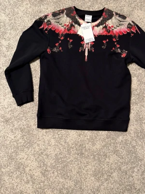 Svart sweatshirt från Marcelo Burlon - Cool svart sweatshirt från Marcelo Burlon med unikt vingmönster i rött, grått och beige över axlar och bröst. Tröjan har rund halsringning och långa ärmar. Perfekt för dig som vill sticka ut med en statement-piece.