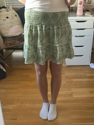 Grön blommig kortkjol med volang - Söt grön kortkjol med vitt blommönster och volang nertill. Kjolen har smock i midjan för en bekväm passform och är perfekt för varmare dagar. Lätt och luftig modell som ger en avslappnad och trendig look.
