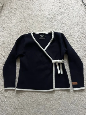 Marinblå omlottkofta  - Snygg marinblå omlottkofta från Holebrook Sweden med vita kanter och knytband i midjan. Perfekt för dig som gillar klassisk och stilren design.  - Mycket bra bra och tjock kvalitetstyg (100% bomull) - Har en knapp och ett resår med flera hål så den sitter bättre och man kan välja hur hårt den ska sitta