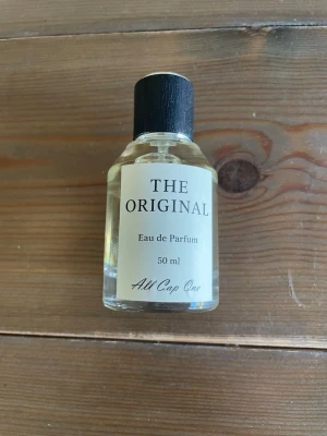 The Original Eau de Parfum 50 ml - Parfymflaska med transparent glas och svart lock. Märkt 'The Original' från All Cap One, 50 ml Eau de Parfum. Flaskan har en stilren, minimalistisk design med silvrig spraypump och vit etikett med svart text.