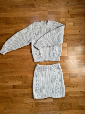 Stickat set i beige med kabelmönster - Snyggt stickat set i beige med tröja och kjol. Båda plaggen har klassiskt kabelstickat mönster och breda ribbade muddar. Perfekt för dig som gillar en mysig och trendig look. Materialet känns mjukt och värmande.