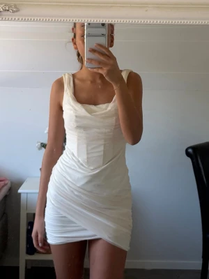 Vit kort korsetklänning med drapering från OhPolly - Snygg vit kort klänning med draperad effekt och breda axelband. Klänningen har en omlottliknande nederdel och en smickrande passform som framhäver figuren.  Klänningen är inte kemtvättad