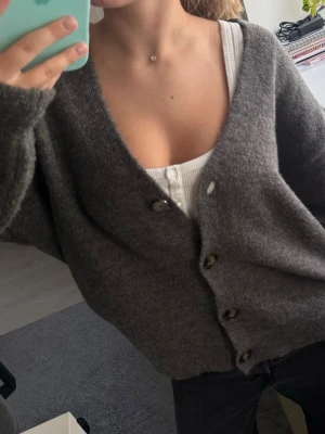 Mörkgrå oversized kofta med knappar - Mysig mörkgrå kofta med v-ringning och stora knappar framtill. Oversized passform och långa ärmar, perfekt att slänga på sig över ett linne eller t-shirt. Koftan är stickad och har en enkel, stilren look som passar till det mesta.