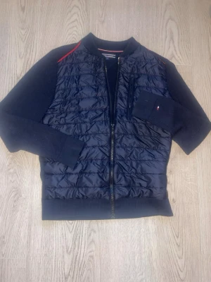 Svart bomberjacka Tommy Hilfiger - Snygg svart bomberjacka från Tommy Hilfiger med quiltad framsida och stickade ärmar. Jackan har dragkedja, ribbade muddar och en diskret logga på ärmen. Röd detalj vid axlarna och bröstficka med dragkedja. Perfekt för en trendig streetstil.