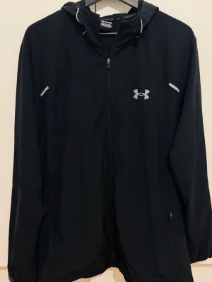 Svart vindjacka från Under Armour - Svart vindjacka från Under Armour med huva och hel dragkedja. Jackan har reflekterande detaljer och logga framtill samt tryckt logotyp på ryggen. Tillverkad i lätt och smidigt material, perfekt för blåsiga dagar. Och lätt regn. 