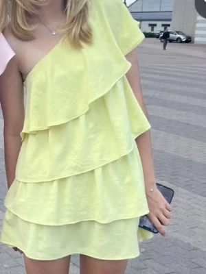 Gul one shoulder volangklänning - Snygg kort gul klänning med one shoulder-design och flera volanglager. Perfekt för sommarens alla tillfällen. Lätt och luftig modell som ger en trendig look.