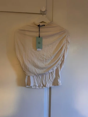 Vit omlottopp från gina tricot - Snygg vit omlottopp från Gina med hög hals och draperad design. Toppen har en unik omlottkänsla och är tillverkad i ett mjukt material som ger en cool och trendig look. Perfekt att styla med jeans eller kjol.