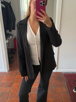 Svart oversized klassisk kavaj - Snygg svart kavaj med enkelknäppning och klassisk krage. Rak passform och två fickor framtill. Perfekt att styla över en topp eller skjorta för en clean look. Materialet ser ut att vara ett slätt tyg, troligen polyester eller bomullsblandning.
