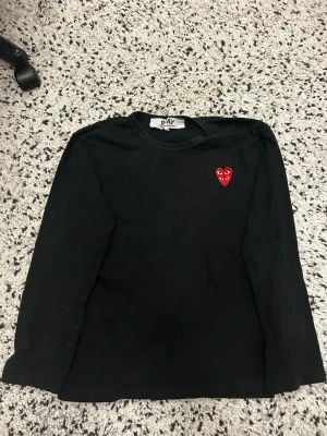 Svart långärmad topp från Comme des Garçons - Svart långärmad topp från Comme des Garçons Play med det ikoniska röda hjärtat med ögon broderat på bröstet. Enkel och stilren design, perfekt för att lyfta din outfit med en cool detalj.