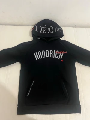 Svart hoodie från Hoodrich - Svart hoodie från Hoodrich med stor vit logga på bröstet och tryck på huvan. Hoodien har magficka och dragsko i halsen. Materialet är mjukt och bekvämt, perfekt för en streetwear-look. Reflekterande tryck på bröstet som syns i ljus.