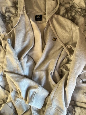 Ljusgrå hoodie från Lyle & Scott - Ljusgrå hoodie från Lyle & Scott med dragkedja, huva med dragsko och fickor framtill. Klassisk gul logga på bröstet. Mjuk bomullsblandning och ribbade muddar vid ärmar och nederkant. Perfekt för en avslappnad och stilren look. (Dragkedjan är av så pris går diskuteras”