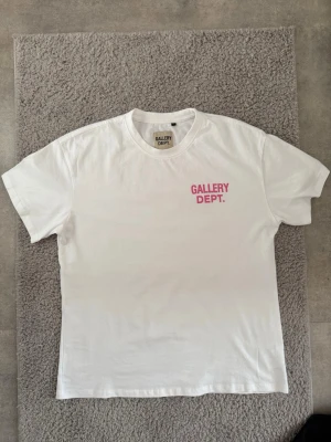 Vit Gallery Dept. t-shirt med tryck - Vit t-shirt från Gallery Dept. med rosa tryck på bröstet och stort tryck på ryggen där det står 'GALLERY DEPT. HOLLYWOOD, CA.'. Klassisk passform och rund halsringning. Perfekt för en clean och trendig streetwear-look.