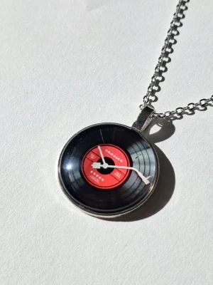 Vinylskive-halsband Crosley - Unikt halsband med ett runt hänge som föreställer en svart LP-SKIVA i 2D med röd etikett och vit tonarm. Hänget har glansig yta och sitter på en silverfärgad kedja. Perfekt för dig som älskar retro och musikvibbar.