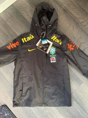 Svart Nike Brazil windbreaker med huva - Svart vindjacka från Nike med Brasil-logga och sponsormärken i gult och orange. Jackan har huva, hel dragkedja och meshfoder på insidan. Perfekt för dig som gillar sportig stil och vill sticka ut med snygga detaljer. Passar främst S.