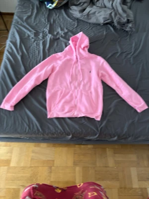 Rosa hoodie från Ralph Lauren - Snygg rosa hoodie från Ralph Lauren med dragkedja framtill och klassisk huva med dragsko. Har två stora fickor på magen och den ikoniska lilla loggan broderad på bröstet. Perfekt för en chill och avslappnad stil.