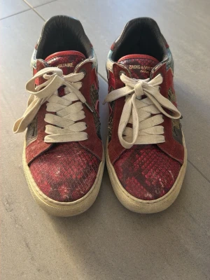 Zadig & Voltaire sneakers med mönster - Unika sneakers från Zadig & Voltaire med röd och svart ormskinnsmönstrad ovandel, detaljer i turkos och grått samt vita snören. Skorna har en låg siluett och vit sula. Perfekt för dig som vill sticka ut med färg och mönster.