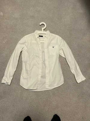 Vit skjorta från Gant med ficka - Klassisk vit skjorta från Gant med bröstficka och broderad logga. Skjortan har knappar hela vägen, button down-krage och lång ärm. Perfekt för en clean och stilren look.