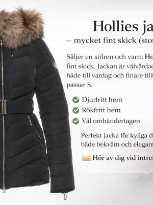 Svart dunjacka med pälskrage Hollies - Snygg svart dunjacka från Hollies med avtagbar pälskrage och markerad midja med bälte. Jackan har dragkedja framtill, två fickor med dragkedja och quiltad design. Perfekt för kalla vinterdagar och ger en trendig look.