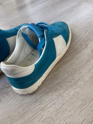 Blåvita sneakers med mockadetaljer - Snygga sneakers i blått och vitt med ovandel i mocka och läder. Skorna har vita detaljer och blå skosnören, samt en klassisk låg siluett. Perfekta för dig som gillar en sportig och stilren look.