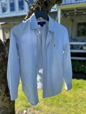 Blårandig skjorta från Ralph Lauren - Klassisk skjorta från Ralph Lauren med tunna blå och vita ränder. Skjortan har lång ärm, knäppning framtill och en liten broderad logga på bröstet. Tillverkad i bomull för en skön och luftig känsla. Perfekt till jeans eller chinos.