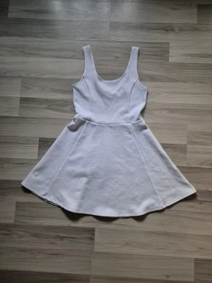 Vit ärmlös kortklänning - Säljer en söt vit kortklänning med rundad halsringning och ärmlös design. Klänningen har en utställd kjol och är tillverkad i ett mjukt, strukturerat tyg som ger en skön känsla. Perfekt för varma dagar och enkel att styla med accessoarer.