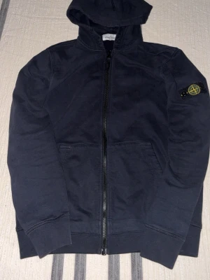 Svart hoodie från Stone Island Junior - Svart hoodie med dragkedja från Stone Island Junior. Klassisk design med huva, två fickor framtill och ikonisk logotyp-patch på vänster ärm. Tillverkad i mjukt bomullsmaterial, perfekt för en avslappnad streetwear-look.