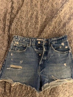 Blåa högmidjade jeansshorts med slitningar - Snygga blå jeansshorts från ZARA med hög midja, slitna detaljer och fransig kant nedtill. Klassisk femficksmodell med knappar framtill. Perfekta för en avslappnad och trendig look.