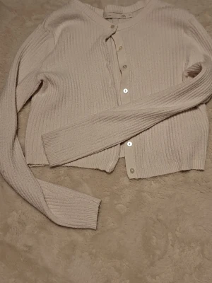 Beige ribbstickad kofta - Mysig beige ribbstickad kofta med rund halsringning och knäppning framtill. Långa ärmar och mjukt material som känns skönt mot huden. Perfekt att slänga på sig över en t-shirt eller linne.