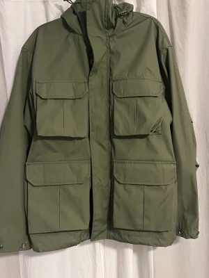 Olivgrön vindjacka från H&M - Stilren olivgrön vindjacka från H&M med fyra stora fickor framtill och huva. Jackan är vattenavvisande och vindtät, perfekt för blåsiga dagar. Praktisk design med dragkedja och tryckknappar. Materialet är syntetiskt och lätt att bära.