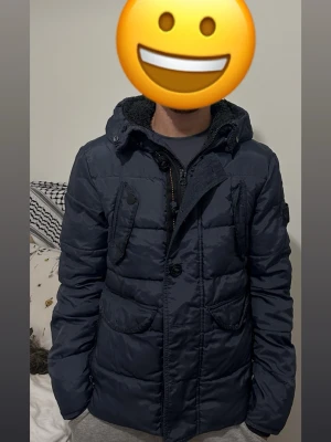 Mörkblå pufferjacka med huva - Snygg mörkblå pufferjacka med huva och dragkedja samt knappar framtill. Jackan har flera stora fickor och en mysig foder på insidan av kragen. Perfekt för kalla dagar och har en modern, avslappnad passform. Jätte fin jacka ingen skador alls men den e liten på mig använt bara en vinter jätte varm och bekväm 