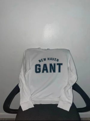 Vit sweatshirt från Gant med tryck - Snygg vit sweatshirt från Gant med stort blått tryck 'NEW HAVEN GANT' på bröstet. Klassisk rund halsringning och ribbade muddar vid ärmslut och nederkant. Perfekt för en avslappnad och stilren look.