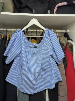 Blå- och vitrandig blus från Monki - Snygg blå- och vitrandig blus från Monki med offshoulder-modell och korta puffärmar. Blusen har knappar framtill och en avslappnad passform. Perfekt för en trendig och luftig look i sommar.