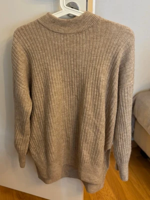 Beige ribbstickad tröja med hög krage - Snygg beige stickad tröja med ribbstickat mönster och högre krage. Tröjan har en avslappnad passform och långa ärmar, perfekt för lager-på-lager. Materialet känns mjukt och skönt mot huden.