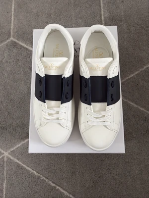 Valentino vita sneakers med svart detalj - Snygga vita sneakers från Valentino Garavani med bred svart läderdetalj över snörningen. Skorna har klassisk låg profil, vita snören och en exklusiv präglad sula. Perfekta för dig som gillar stilrena och lyxiga sneakers.