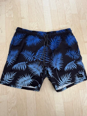 Svarta badbyxor med blått palmbladsmönster - Snygga svarta badbyxor från Aikoon med blått palmbladsmönster. De har elastisk midja med dragsko för skön passform. Perfekta för stranden eller poolen och har en modern, avslappnad stil.