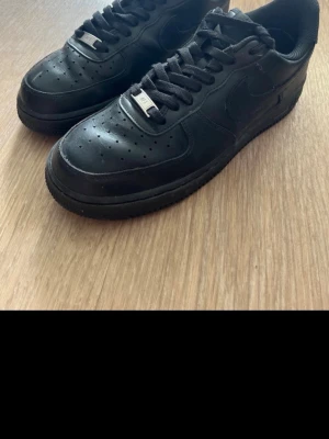 Nike Air Force 1 Low helsvart läder - Klassiska Nike Air Force 1 Low i helsvart läder med svart sula och svarta skosnören. Ikonisk siluett med perforerad tåbox och metall-detalj på snörningen. Perfekta sneakers för dig som gillar en clean och tidlös look.